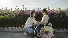 Symbolfoto: Mutter kniet vor ihrer Tochter, die im Rollstuhl sitzt, hält ihre Hand. Im Hintergrund eine Blumenwiese