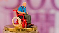 Rollstuhl-Figürchen auf einem Münzstapel, im Hintergrund ein 500-Euro-Schein