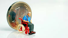 Symbolfoto: Euromünze steht auf ihrem Rand; davor ein Figürchen eines älteren Herrn im Rollstuhl