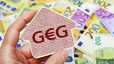 Hand hält Schild mit der Aufschrift "GEG". Dahinter liegen viele Euro-Scheine.