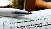 Symbolfoto: Ein Formular einer Steuererklärung, im Hintergrund Münz-Stapel und ein Kugelschreiber