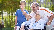 Symbolfoto: Drei Generationen stehen zusammen und lachen - Enkelsohn, Vater und Großmutter, die im Rollstuhl sitzt