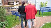 Symbolfoto: Eine ältere Frau mit einem Gehstock, sie ist bei einer jüngeren Frau untergehakt. Beide sieht man nur von hinten. Sie gehen auf einem Weg durch eine Grünanlage.