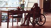 Symbolfoto: Eine alte Dame im Rollstuhl sitzt an einem Tisch, den Kopf auf eine Hand gestützt. Die Situation wirkt trist und düster.