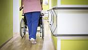 Symbolfoto: Ein Gang in einem Krankenhaus oder Pflegeheim. Eine Pflegerin ist von hinten zu sehen, sie schiebt einen Rollstuhl.