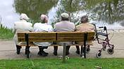 Symbolfoto: Seniorinnen und Senioren sitzen auf einer Parkbank, man sieht ihre Rücken. Neben der Bank steht ein Rollator.
