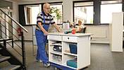 Symbolfoto: Älterer Herr im Blaumann rollt einen Arbeitstisch mit Werkzeug durch ein Büro