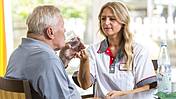 Eine Pflegerin reicht einem älteren Mann ein Glas mit Wasser.