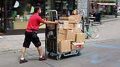 Das Bild zeigt einen Paketboten mit einem Rollwagen voller Pakete durch eine Fußgängerzone eilen.