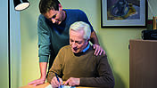 Das Bild zeigt einen jüngeren und einen älteren Mann. Dieser schreibt auf einem Dokument.
