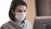Das Bild zeigt eine Frau mit Mundschutzmaske vor dem Computer