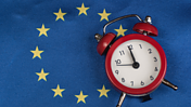 Das Bild zeigt im Hintergrund als HIntergrund das Design der EU-Flagge mit Blau und Sternen. Im Vordergrund ist eine Uhr zu sehen, die kurz vor 12 steht.
