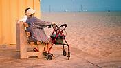 Zwei ältere Personen sitzen auf einer Bank, davor Strand, eine hat einen Rollator