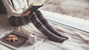 Eine junge Frau liest ein Buch, sie trägt warme Wollsocken und neben ihr steht ein tablett mit Plätzchen