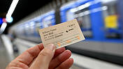 Eine Hand hält ein 9-Euro-Ticket, im Hintergrund steht eine Bahn