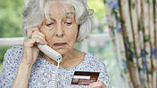Seniorin hält Telefonhörer in der Hand und schaut besorgt auf eine Kreditkarte.
