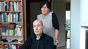 Das Ehepaar Gabriele Mair-Bolland und Klaus Bolland in seiner Wohnung. Er sitzt im Rollstuhl, sie steht hinter ihm und hält die Griffe des Rollstuhls.