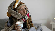 Junge Frau sitzt im Bett mit dickem Schal und Mütze, in der Hand hält sie einen Becher mit Tee.