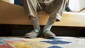 Symbolfoto: älterer Mann, der Pantoffel trägt, sitzt auf dem Rand eines Pflegebettes.