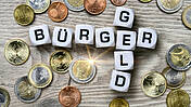 Symbolfoto: Verschiedene Euro- und Centmünzen auf einer Tischplatte, darauf liegen Buchstabenwürfel, die wie beim Scrabble-Spiel das Wort "Bürgergeld" bilden