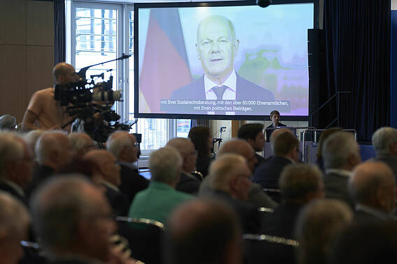 Blick in den Tagungssaal, auf einer großen Leinwand wird die Videobotschaft von Bundeskanzler Olaf Scholz gezeigt.