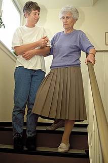 Symbolfoto: Eine jüngere Frau hilft einer Seniorin, eine Treppe hinabzugehen. Sie hält die ältere Frau am Arm eingehakt fest.