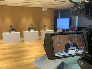 Blick aufs Podium bei der VdK-Veranstaltung, durch eine Kamera sieht man Prof. Andreas Büscher, der gerade vorträgt.