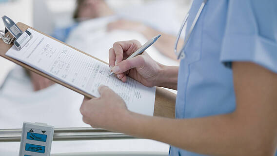 Krankenschwester notiert Informationen auf einem Klemmbrett, Patient im Hintergrund