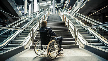 Geschäftsmann in einem Rollstuhl, umgeben von Treppen und Rolltreppen