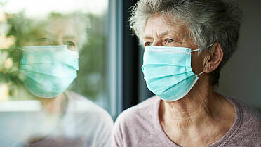 Eine alte Frau trägt eine OP-Maske und schaut aus dem Fenster