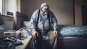 Älterer Mann sitzt zu Hause auf seinem Bett. Er trägt ein medizinisches Beatmungsgerät über seinem Gesicht, das an einer Maschine neben ihm befestigt ist.