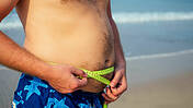 Mann in Badehose misst seinen Bauchumfang am Strand mit einem Schneidermassband