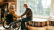 Älteres Ehepaar, Frau im Rollstuhl, Mann sitzt neben ihr auf großer Fensterbank und schaut sie an