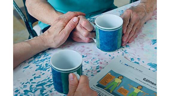 Das Bild zeigt eine ältere Frau und einen Mann. Sie sitzen an einem Küchentisch und halten jeweils eine Tasse in der Hand. Der mann drückt die Hand der Frau. Vor ihnen liegt die VdK-Zeitung.
