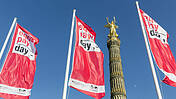 Das Bild zeigt drei Flaggen mit dem Logo des Equal-Pay-Days. Im Hintergrund sieht man die Siegessäule in München.