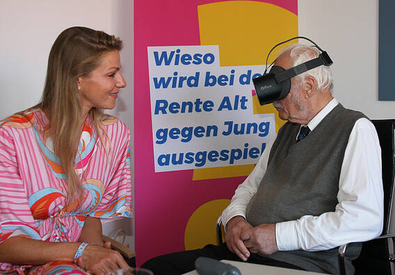 Auf dem Foto sieht man den VdK-Ehrenamtlichen Joachim Dietrich (rechts), der eine VR-Brille trägt. Links neben ihm sitzt Carolina Bendlin, Geschäftsführerin von Granny Vision, die ihm den Umgang mit der VR-Brille erklärt.