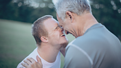 Das Bild zeigt einen lächelnden jungen Mann mit Downsyndrom Stirn an Stirn mit einem liebevollen älteren Herrn auf einer Wiese.
