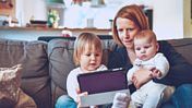 Das Bild zeigt eine Mutter mit zwei Kleinkindern auf dem Schoß. Sie sitzen anscheinend im Wohnzimmer und Blicken zu dritt auf ein Tablet.