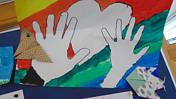 Das Foto zeigt den Ausschnitt einer Pinnwand u.a. mit angehefteten Origami Basteleien, einem bunten Bild, auf dem zwei Hände auf einem Herzen gemalt sind.