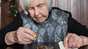 Das Bild zeigt eine ältere Frau zu Hause am Tisch sitzend, sie leert ihre Geldbörse vor sich aus und zählt die Münzen.