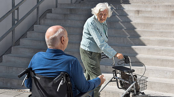 Symbolbild: Eine Frau mit Rollator und ein Rollstuhlfahrer vor einer Treppen.