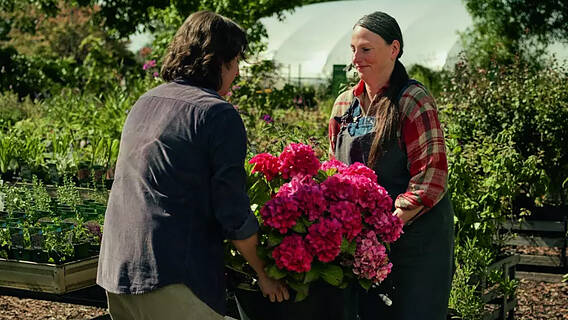 Ein Bild aus dem DRV-Werbespot "Reha hat ein Zuhause". Ein Mann und eine Frau in einer Gärtnerei tragen gemeinsam einen schweren Blumenkübel mit Hortensien.'