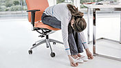 Ein Frau, sitzend auf einen Bürostuhl, beugt sich bis zum Boden.