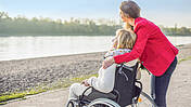 Ein Frau im Rollstuhl und eine Begleitperson verweilen vor einem Fluss.