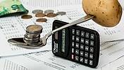 Ein Löffel auf einen hochkant gestellten Taschenrechner als Waage zwischen Münzen und einer Kartoffel.