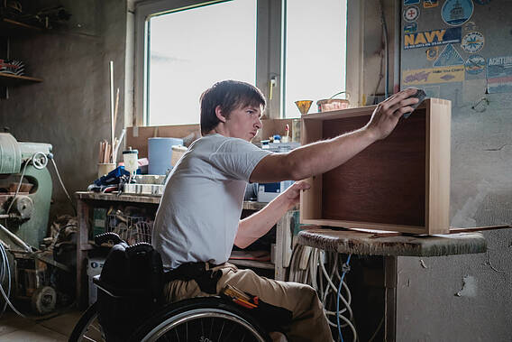 Foto eines Jungen Mannes, der als Tischler an einer Schublade arbeitet und im Rollstuhl sitzt. 