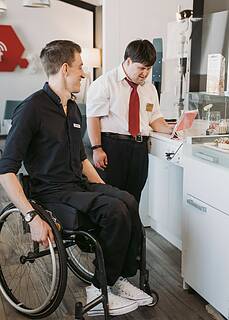 Foto von einem Mann im Rollstuhl und einem Mann mit Down-Syndrom bei der Arbeit in einem Café