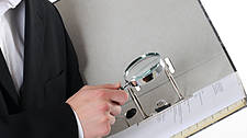 Symbolfoto: Ein Mann in einer Richterrobe schlägt etwas in einem Aktenordner nach, in der Hand eine Lupe