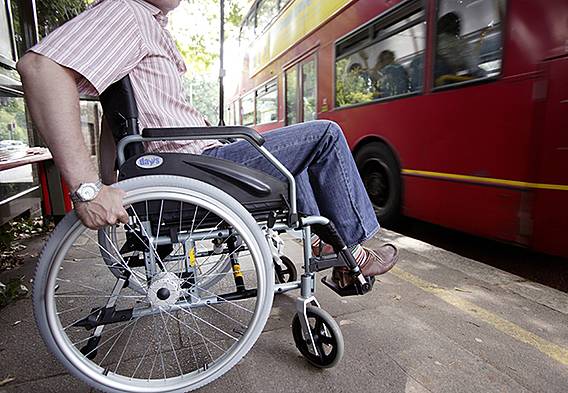 Symbolfoto: Mann im Rollstuhl auf dem Bürgersteig, ein Bus fährt vorbei