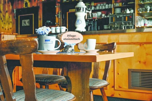 Man sieht den Innenraum einer Wirtschaft. Im Vordergrund sind zwei St&uuml;hle und ein Tisch. Auf dem Tisch stehen zwei Tassen und ein Schild mit "Pflegestammtisch".